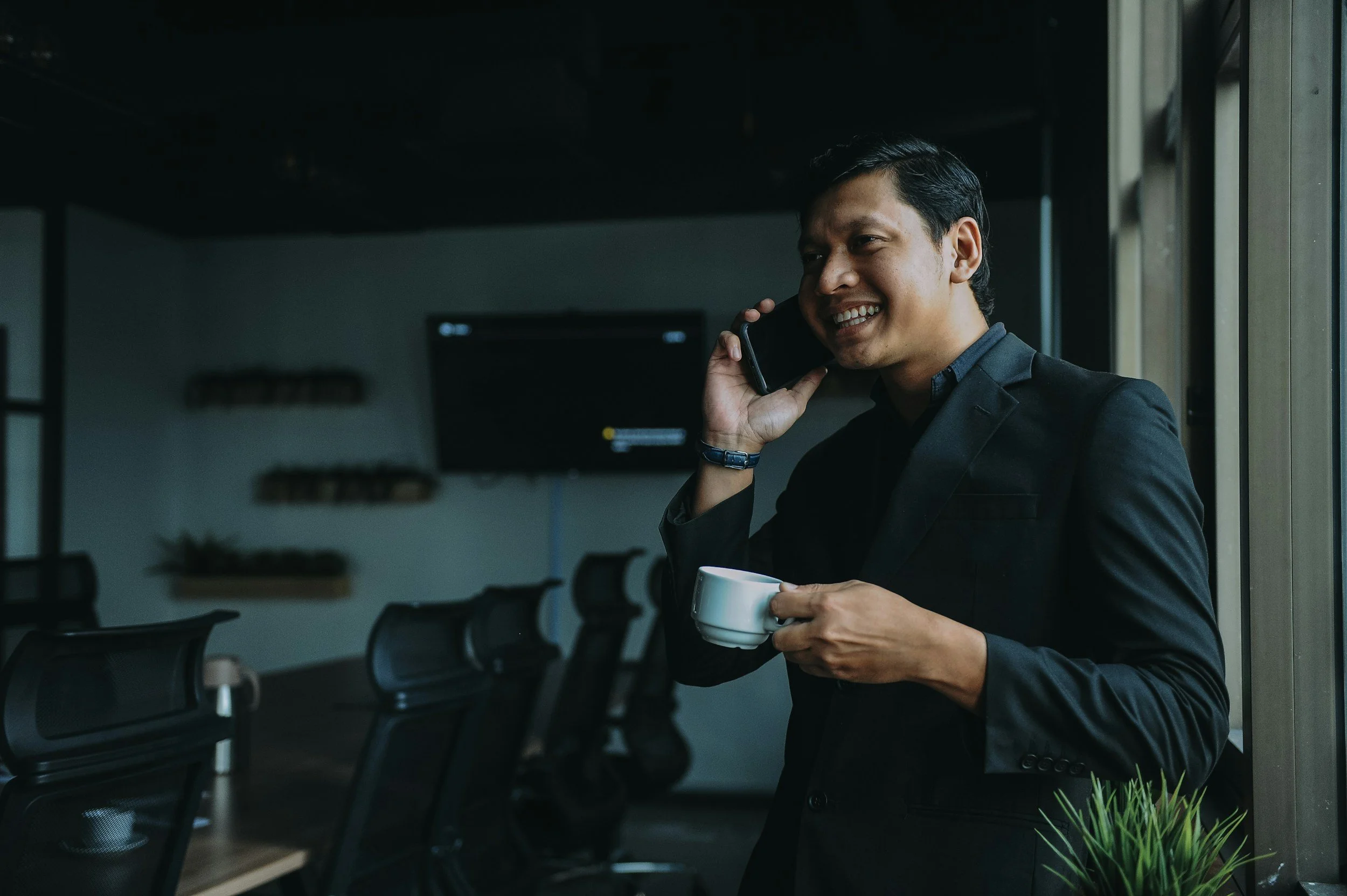 A smiling man in a dark suit talking on a phone while holding a white coffee cup in a modern office.