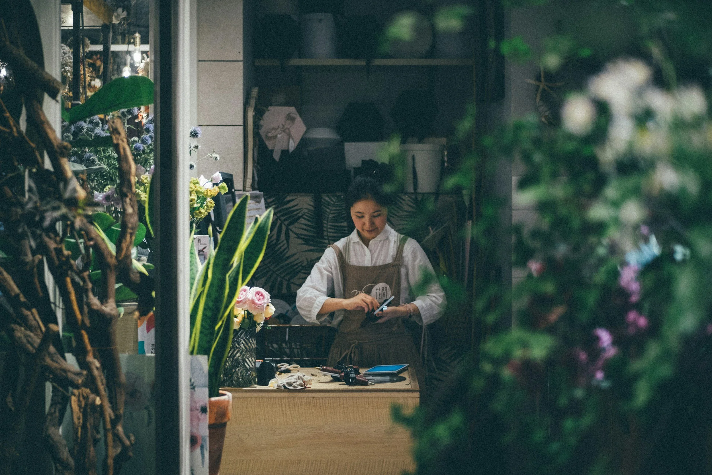 A young woman in a white shirt and apron is standing behind a wooden counter inside a flower shop, using her phone, surrounded by potted plants and flowers.