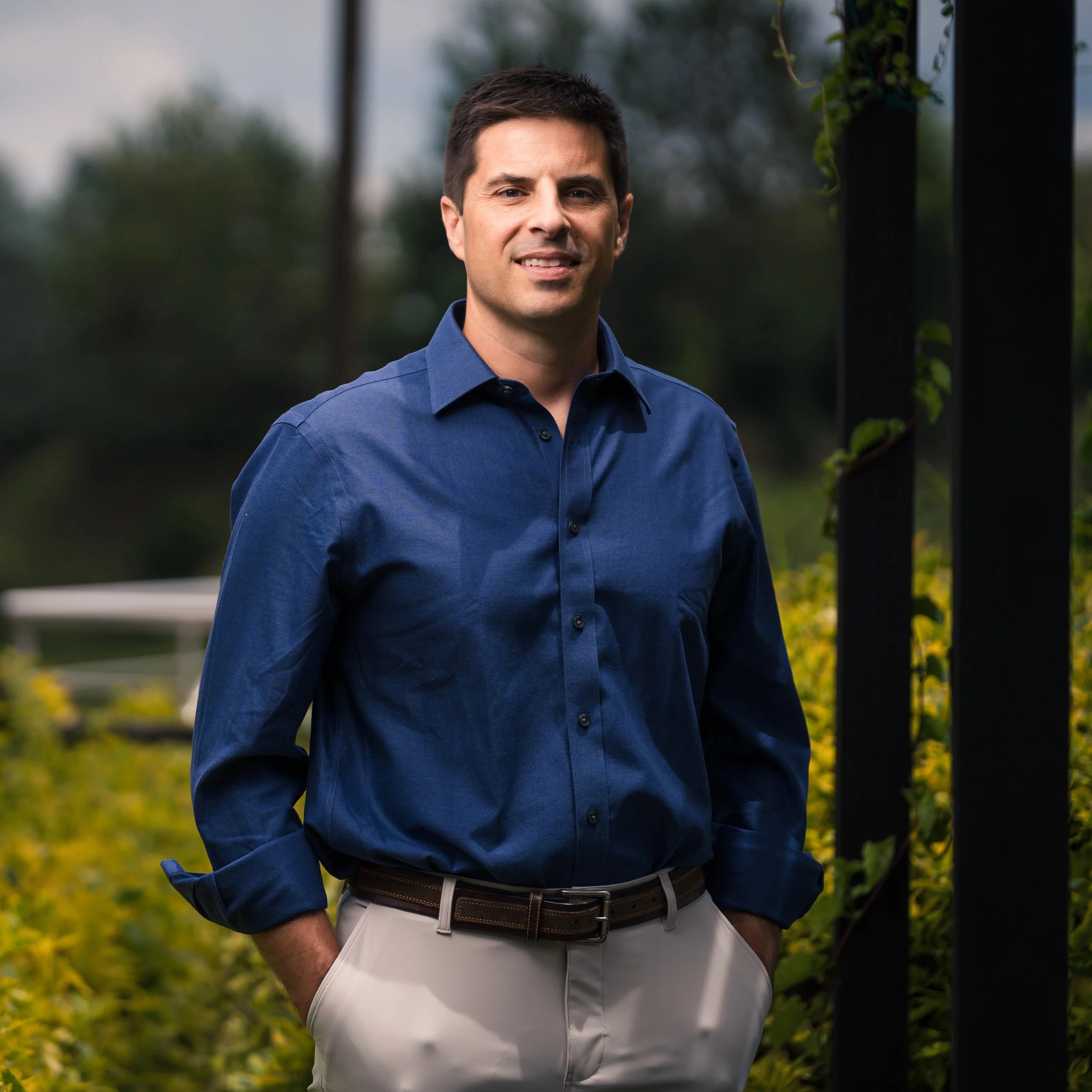 A man standing outdoors with trees and greenery in the background, wearing a blue button-up shirt and beige pants.