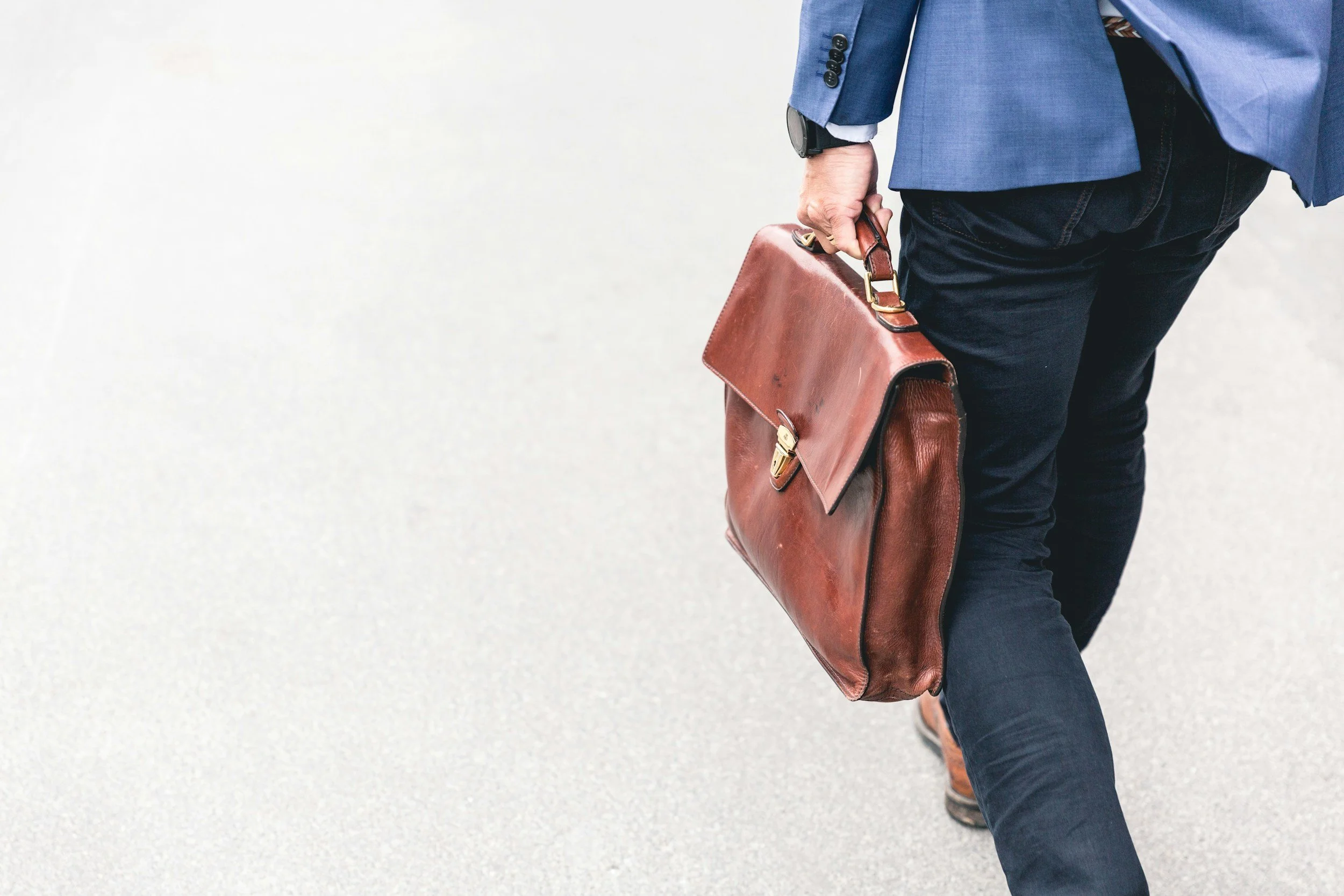A person in business attire carrying a brown leather briefcase, walking on a light-colored surface.