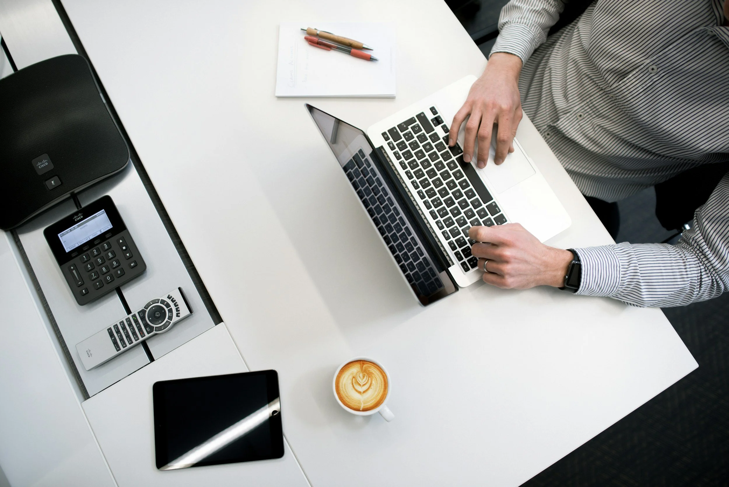 Overhead view of a person working on a laptop at a white desk, with a coffee cup, tablet, phone, and remote control nearby.