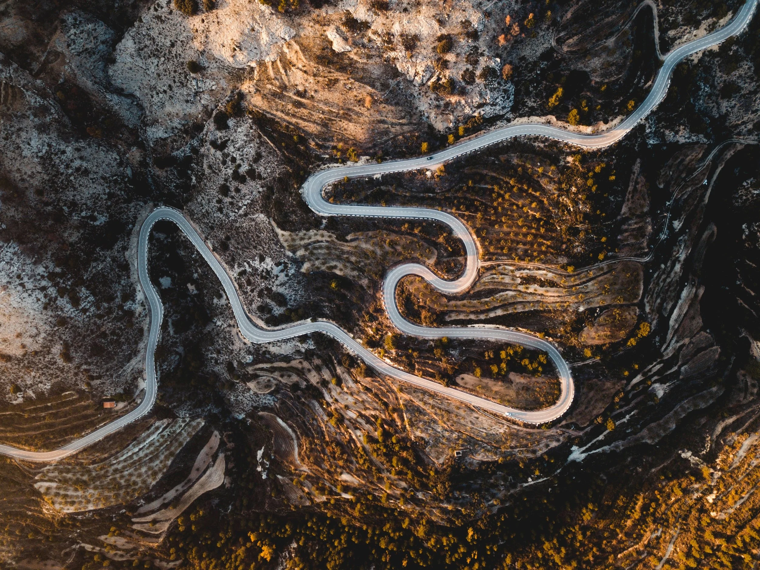 An aerial view of a winding mountain road through a rugged landscape with sparse vegetation and patches of trees.