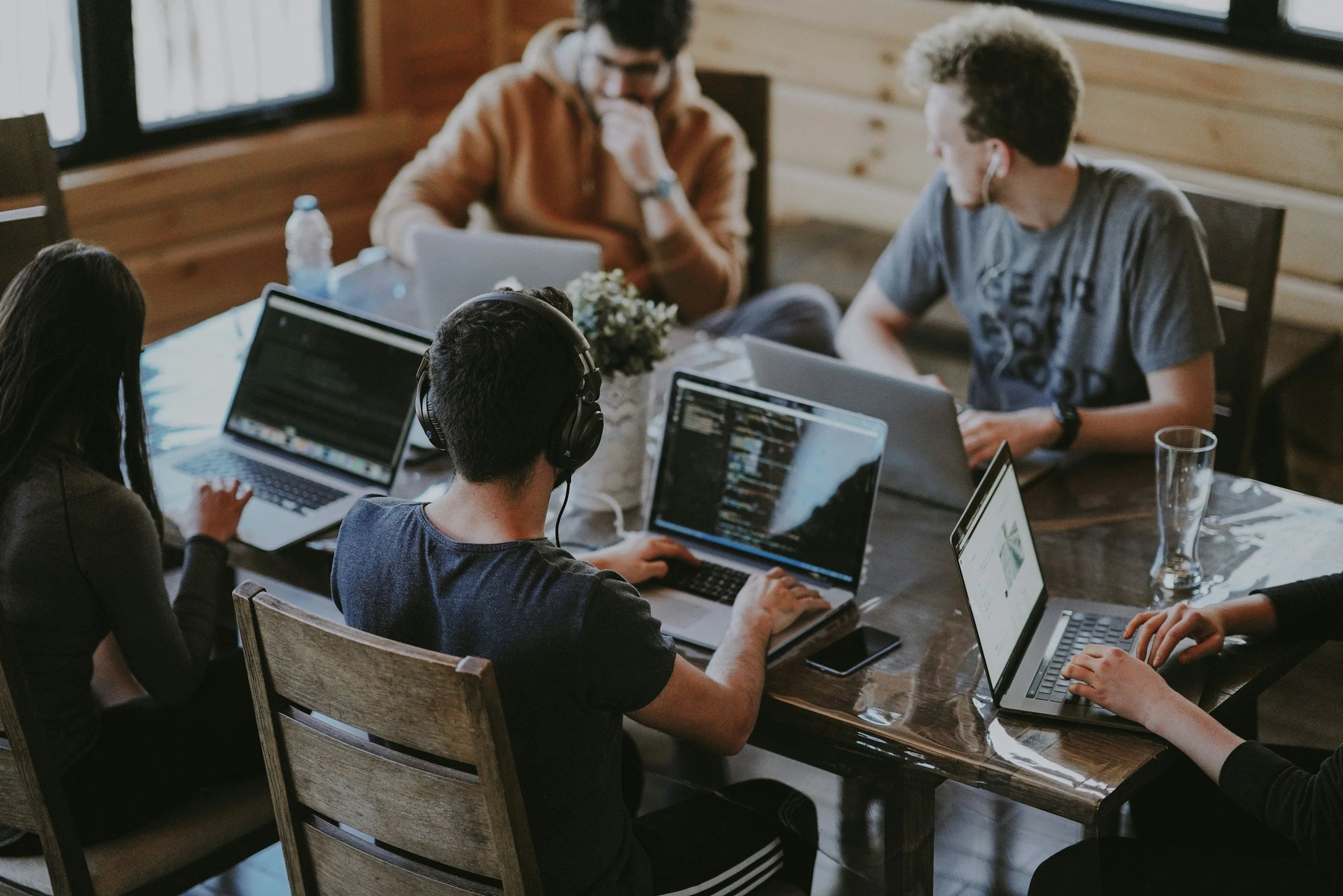 A group of five young adults working on laptops at a wooden table in a cozy room with wooden walls and large windows, some wearing headphones and concentrating on their screens.