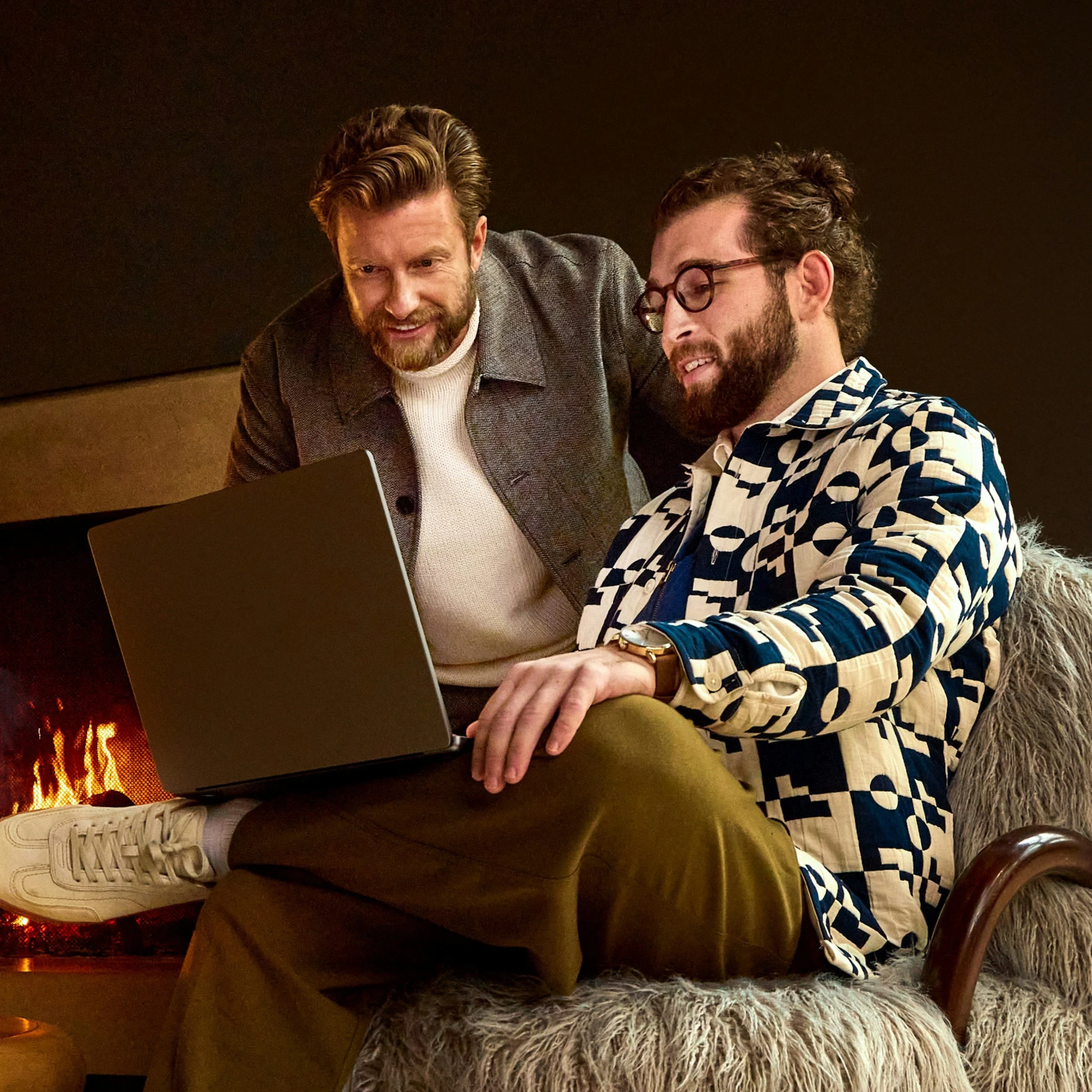 Two men sitting by a fireplace, looking at a laptop screen, one with glasses and patterned blazer, the other with a beard and gray jacket. They are in a cozy, warmly lit room.