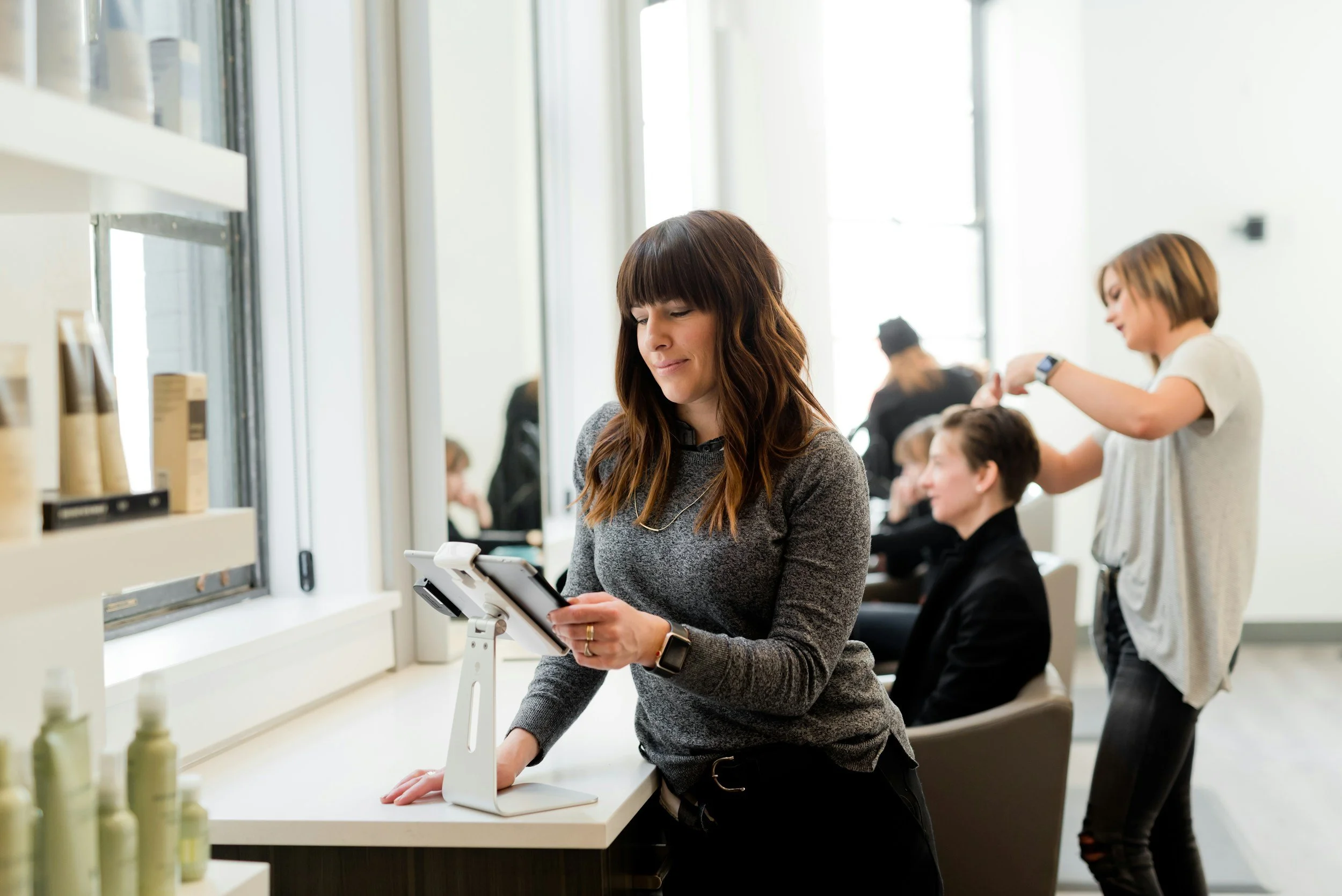 A woman at a salon or spa, using a tablet, while a hairstylist works on another customer's hair in the background.