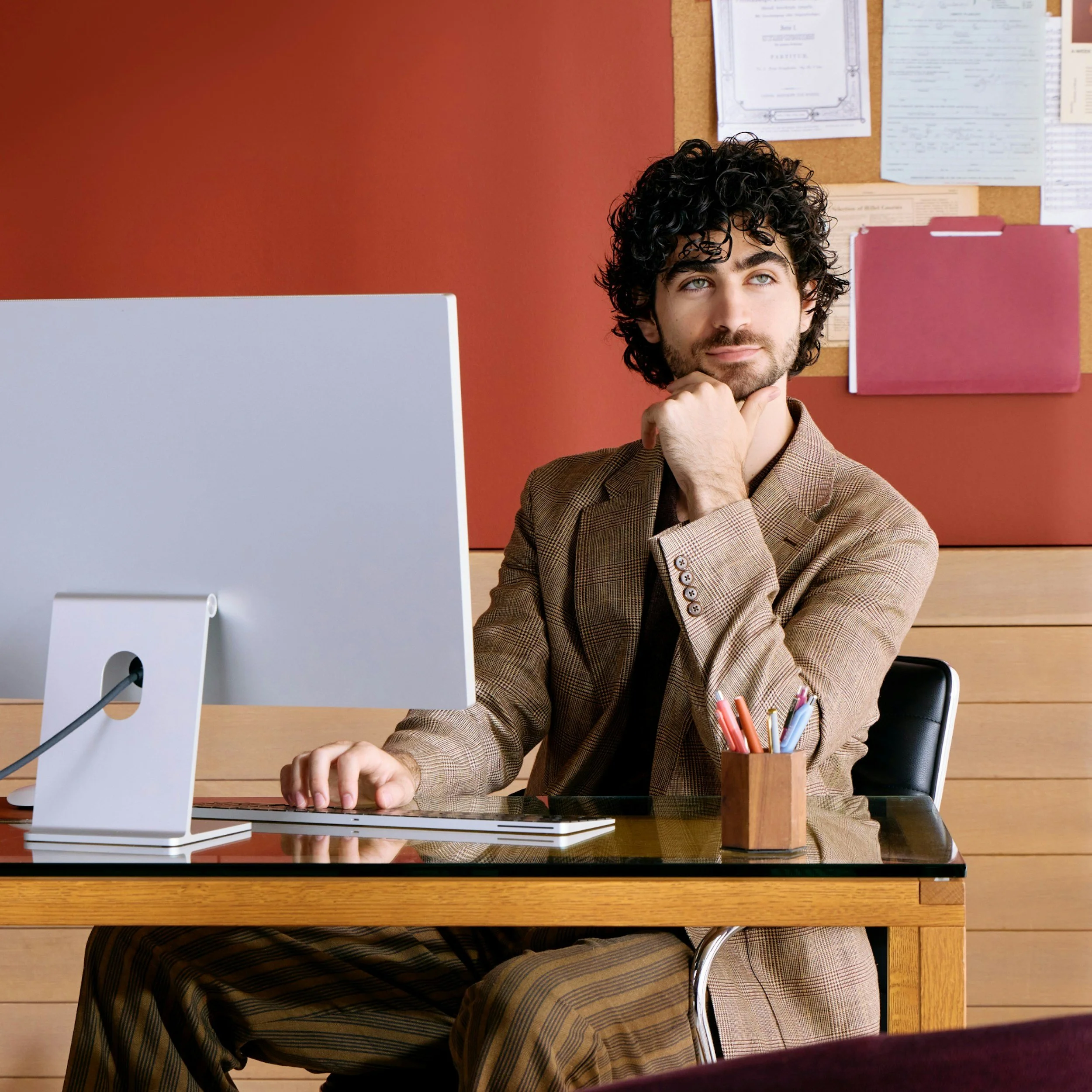 A man with dark, curly hair and a beard sits at a wooden desk in an office, wearing a brown checked suit. He appears thoughtful, resting his chin on his hand. There is a computer monitor, a small wooden cup with colorful pens, and a keyboard on the desk. Behind him is a corkboard with various papers and a pink file folder pinned to the wall.