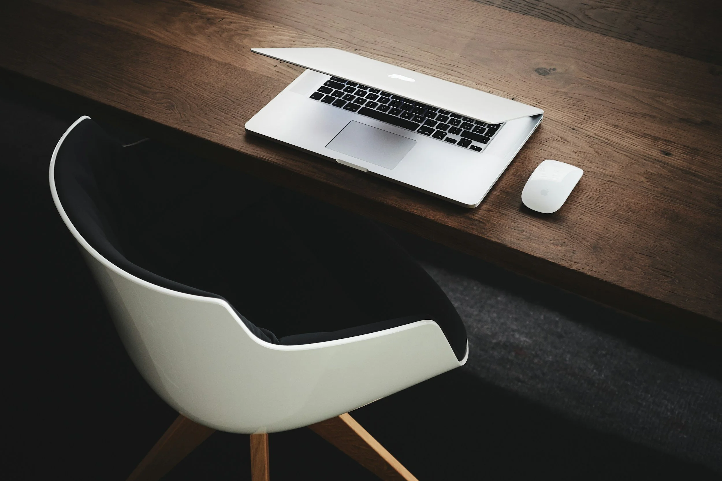 A wooden desk with a silver MacBook laptop partly open and a white Apple Magic Mouse beside it, with a black and white modern chair in front of the desk.