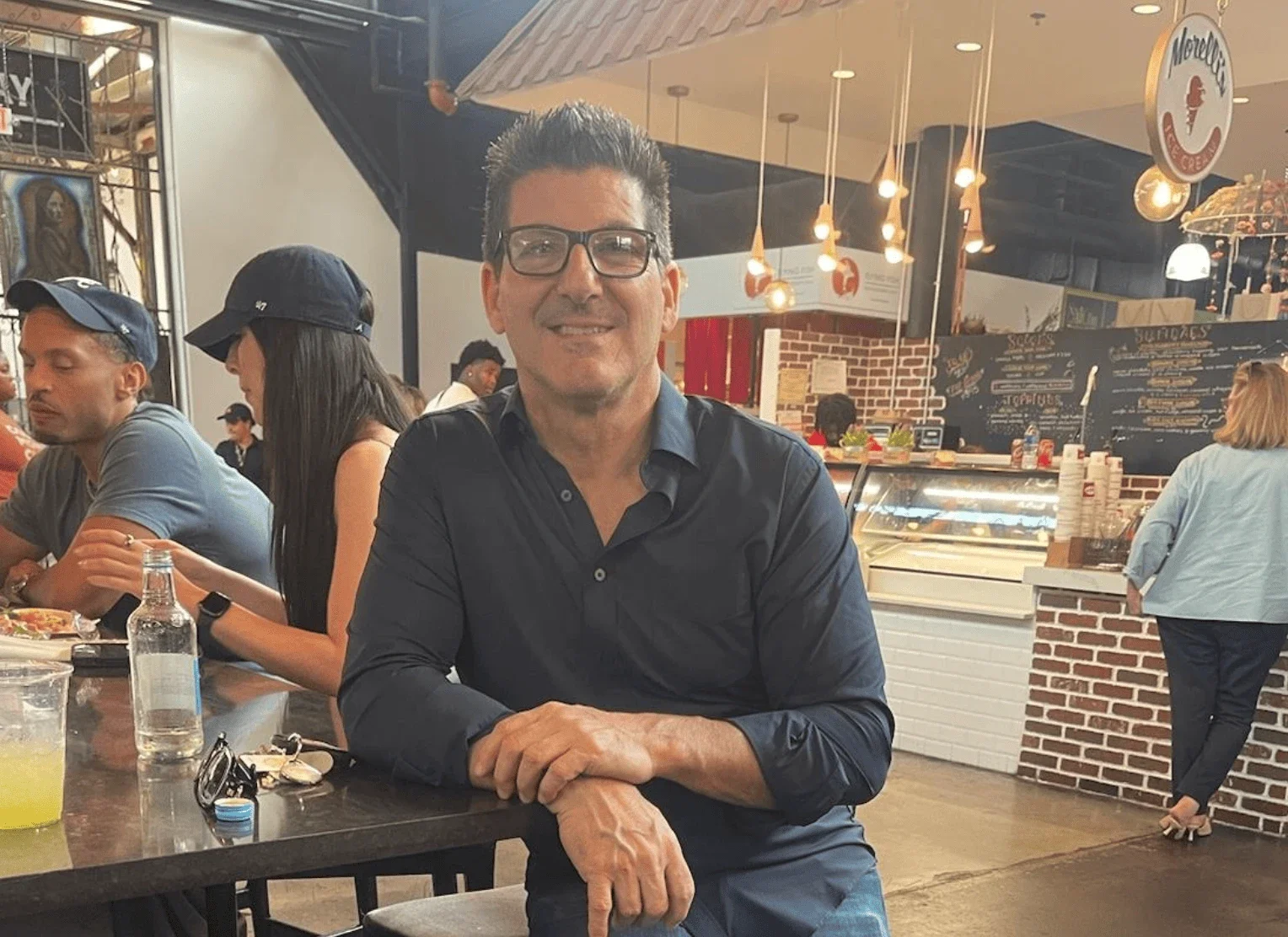 A man with glasses and dark hair smiling at a restaurant or cafe, sitting with his arms crossed on a table.